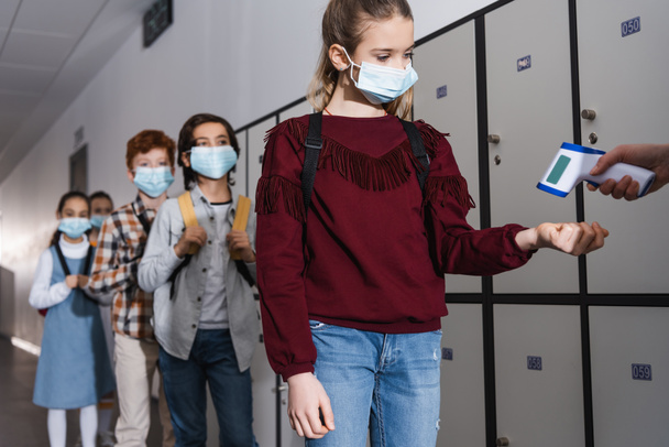 Mädchen in medizinischer Maske misst Temperatur in der Nähe von Lehrerin mit kontaktlosem Thermometer und Klassenkameraden in der Schule - Foto, Bild