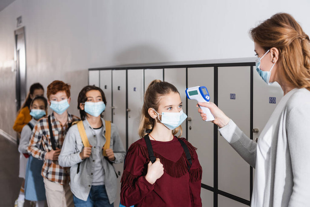 Tanár ellenőrzése hőmérséklet a diákok orvosi maszkok érintésmentes hőmérő a folyosón   - Fotó, kép