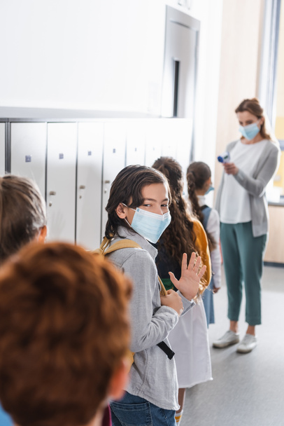Boy in medical mask waving hand near classmates and teacher with infrared thermometer on blurred background  - Photo, Image