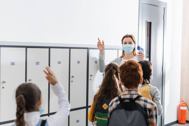 Lehrer mit medizinischer Maske hält kontaktloses Thermometer in der Nähe von Schülern auf verschwommenem Vordergrund im Flur  - Foto, Bild
