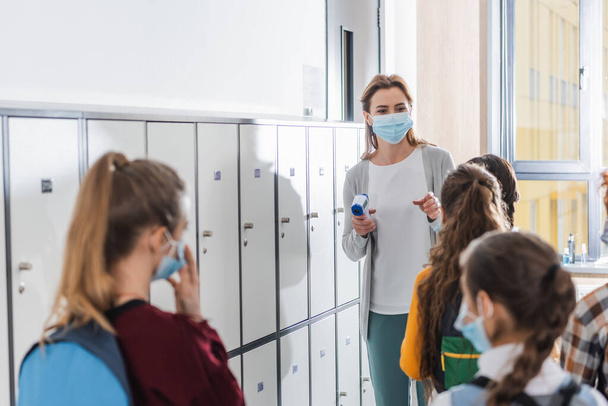 Insegnante con termometro a infrarossi che punta con il dito ai bambini in maschere mediche in primo piano sfocato a scuola - Foto, immagini