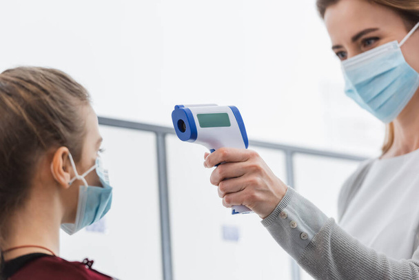 Berührungsloses Thermometer in der Hand des Lehrers in medizinischer Maske zur Temperaturmessung der Schülerin auf verschwommenem Vordergrund  - Foto, Bild