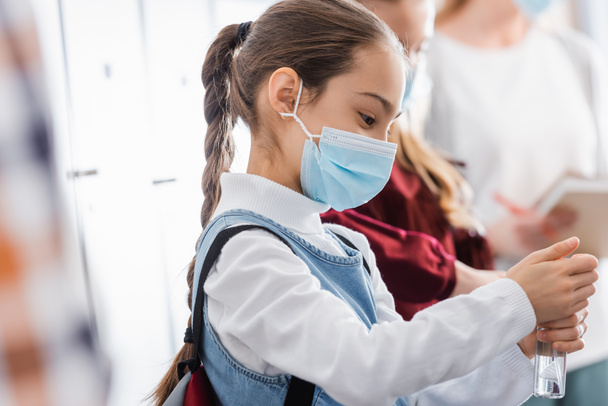 Schoolkid in protective mask using hand sanitizer near pupil on blurred background  - Photo, Image