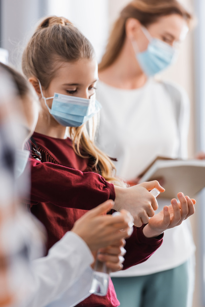 Studentessa in maschera medica spruzzando disinfettante mano vicino insegnante su sfondo sfocato  - Foto, immagini