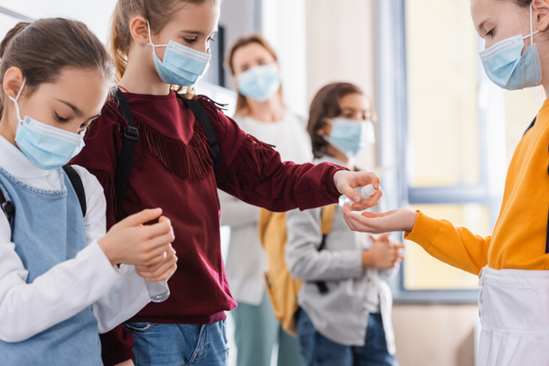 Studenti in maschere mediche utilizzando disinfettante mano vicino insegnante su sfondo sfocato  - Foto, immagini