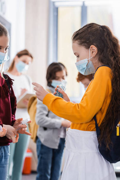 Pupils in medical masks holding disinfector near classmates and teacher on blurred background  - Photo, Image