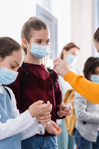 Un écolier portant un masque médical tenant un désinfecteur près de ses amis au premier plan flou à l'école  - Photo, image