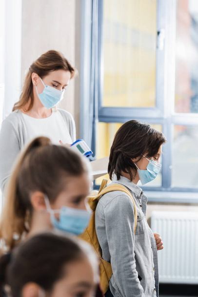 Iskolásfiú orvosi maszkban áll a tanár közelében infravörös hőmérővel és osztálytársaival  - Fotó, kép
