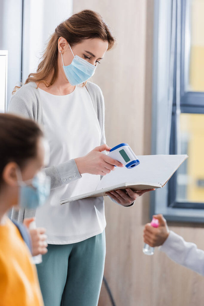 Teacher in medical mask holding infrared thermometer and notebook near pupils on blurred foreground  - Photo, Image