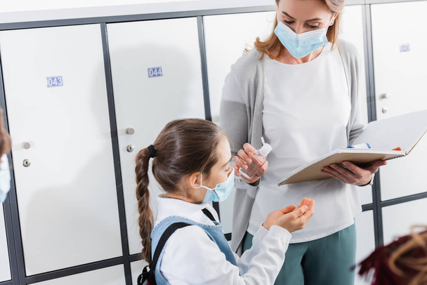 Insegnante con notebook che tiene disinfettante vicino a scolaro in maschera medica in sala - Foto, immagini