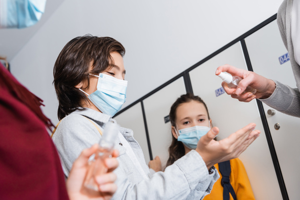 Ragazzo in maschera protettiva in piedi vicino insegnante con disinfettante e amici su sfondo sfocato in sala  - Foto, immagini