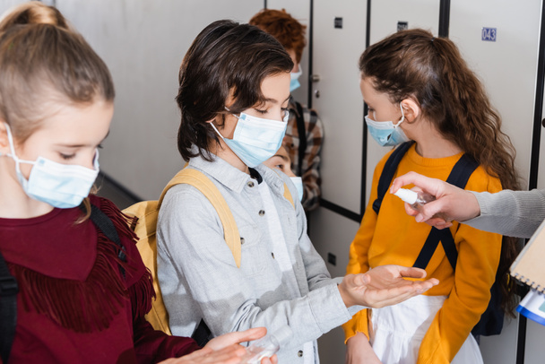 Teacher holding hand sanitizer near pupils in medical masks in school hall  - Photo, Image