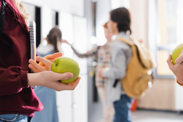 Okul koridorundaki bulanık arka planda sınıf arkadaşlarının yanında elma ve defter tutan kız.  - Fotoğraf, Görsel
