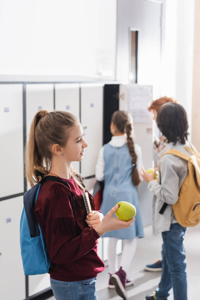 Веселый школьник с зеленым яблоком и блокнотом возле учеников на размытом фоне в зале  - Фото, изображение