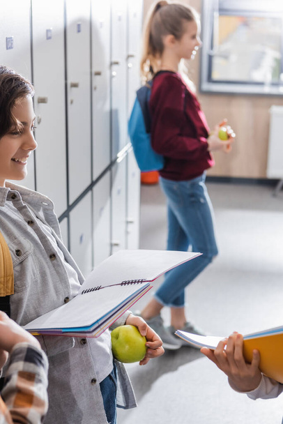 Studentessa sorridente con mela e taccuino in piedi vicino agli armadietti e compagni di classe in primo piano sfocato  - Foto, immagini