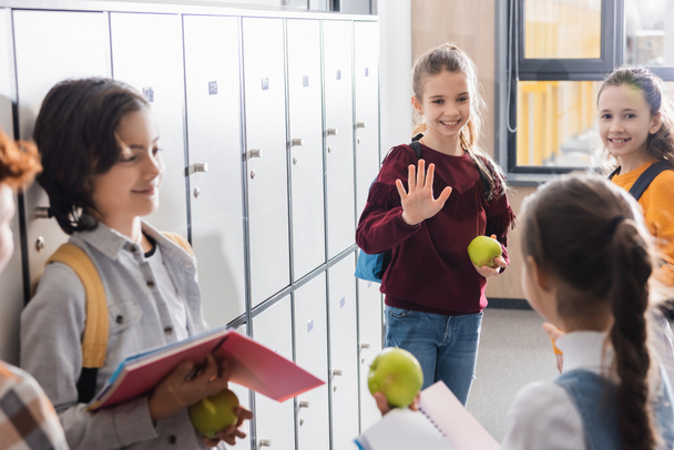 Усміхнена школярка з яблуком махає рукою однокласникам в шкільному коридорі
  - Фото, зображення