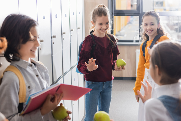 Schoolmeisje met appel en rugzak zwaaien hand naar vrienden op wazig voorgrond in school hal  - Foto, afbeelding