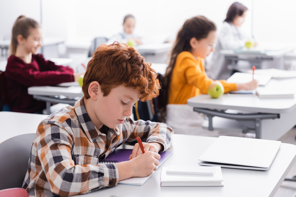 Μαθητής γράφει σε σημειωματάριο κοντά σε φορητό υπολογιστή και smartphone στο γραφείο στην τάξη  - Φωτογραφία, εικόνα
