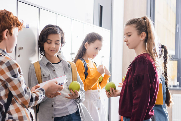 Studentessa con mela fresca in piedi vicino agli amici con taccuino e zaini nel salone della scuola  - Foto, immagini