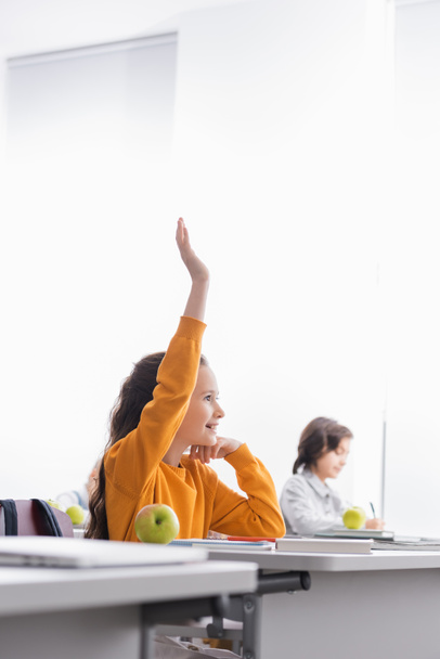 Улыбающийся ребенок с поднятой рукой сидит рядом с яблоком и книги на столе во время урока  - Фото, изображение
