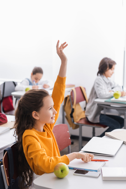 Sonriente niño levantando la mano cerca de dispositivos, manzana y cuaderno en el escritorio de la escuela  - Foto, imagen