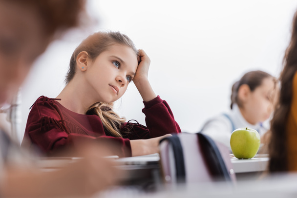 Школьница смотрит в сторону рядом с зеленым яблоком на столе и одноклассники на размытом переднем плане  - Фото, изображение
