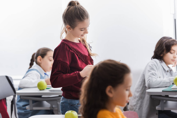 Lächelndes Kind neben Apfel auf Schreibtisch und Mitschüler auf verschwommenem Hintergrund in der Schule  - Foto, Bild
