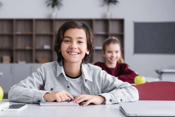 Улыбающийся ребенок смотрит на камеру рядом с устройствами и ноутбуком на столе в классе  - Фото, изображение