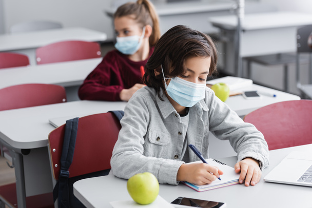Школярка в медичній масці пише на блокноті біля гаджета і яблука на розмитому передньому плані
  - Фото, зображення