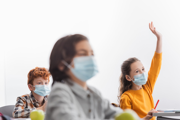 Colegiala en máscara médica levantando la mano cerca de compañeros de clase en clase  - Foto, imagen