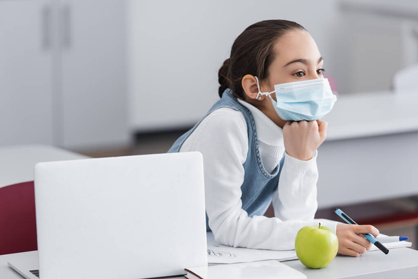 Μαθήτρια με ιατρική μάσκα που κρατάει στυλό κοντά σε μήλο και φορητό υπολογιστή στο γραφείο  - Φωτογραφία, εικόνα