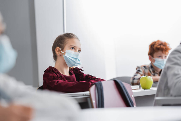 Écolier en masque de protection assis près de pomme mûre sur le bureau et camarades de classe sur fond flou  - Photo, image