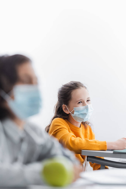 κορίτσι με ιατρική μάσκα κοντά συμμαθητή σε θολή πρώτο πλάνο - Φωτογραφία, εικόνα