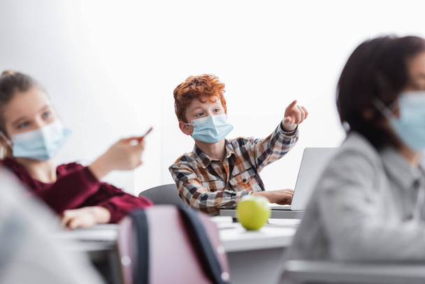 tanulók orvosi maszkban, ujjal és tollal mutogatva az iskolában, homályos előtér - Fotó, kép