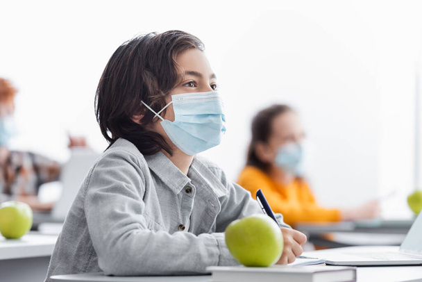 concentrated schoolboy in medical mask writing during lesson near classmates on blurred background - Photo, Image