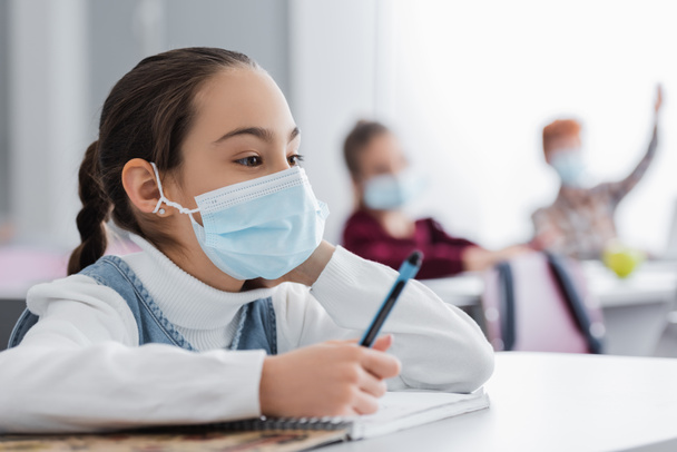 Schulkind mit medizinischer Maske hält Stift während des Unterrichts in der Nähe von Klassenkameraden auf verschwommenem Hintergrund - Foto, Bild