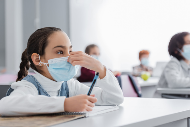 μαθήτρια αγγίζοντας ιατρική μάσκα κρατώντας στυλό κατά τη διάρκεια του μαθήματος - Φωτογραφία, εικόνα