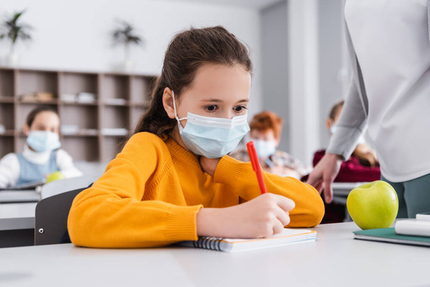 školačka v lékařské masky psaní v notebooku v blízkosti učitel a spolužáci na rozmazaném pozadí - Fotografie, Obrázek