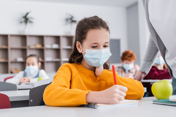 školačka v lékařské masce psaní v notebooku v blízkosti jablka a učitel ve třídě - Fotografie, Obrázek