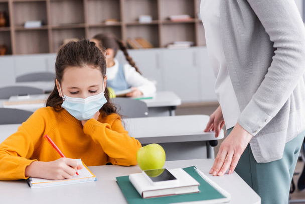 σχολιαρόπαιδο που γράφει στο σημειωματάριο κοντά στο μήλο, smartphone και δάσκαλος στην τάξη - Φωτογραφία, εικόνα