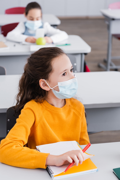 Tıp maskeli kız sınıfta defterin yanından uzağa bakıyor. - Fotoğraf, Görsel