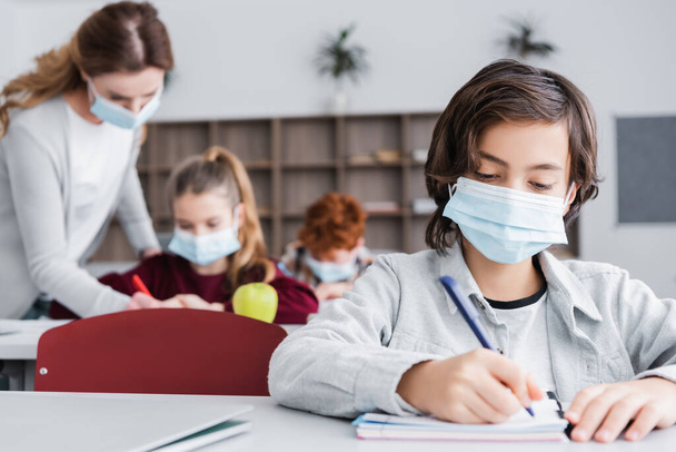 schoolboy in medical mask writing in notebook near classmates and teacher on blurred background - Photo, Image
