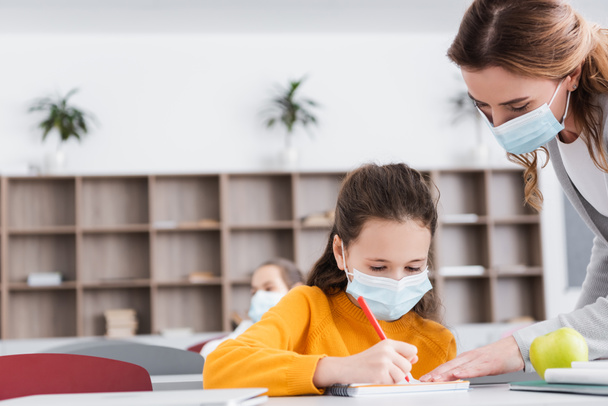 учитель в медицинской маске возле школьницы, которая пишет в блокноте во время урока - Фото, изображение
