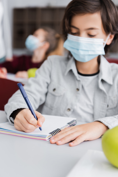 schooljongen in medisch masker schrijven in notebook op wazig achtergrond - Foto, afbeelding