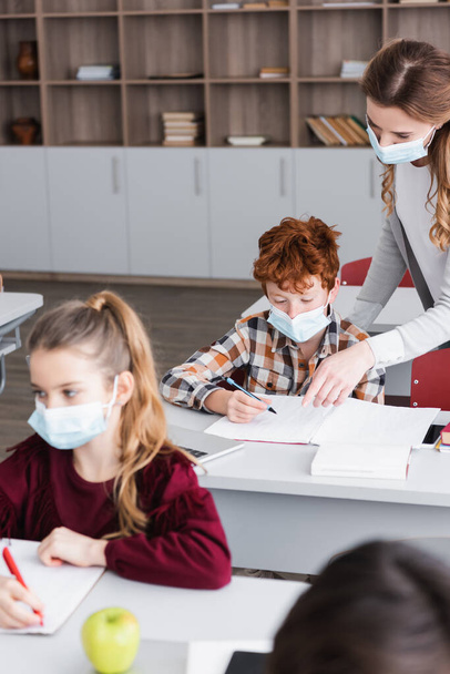 teacher pointing with finger near redhead boy in medical mask writing in notebook during lesson, blurred foreground - Photo, Image