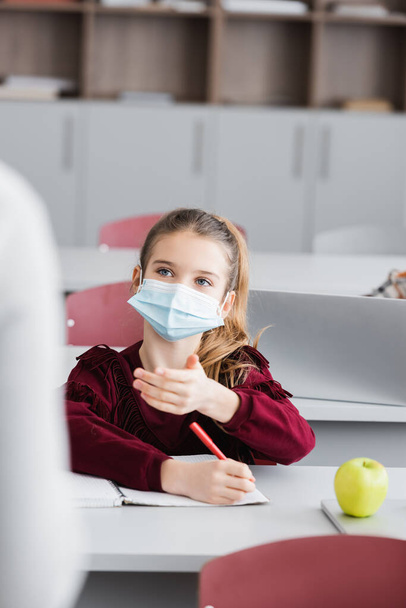 colegiala en máscara médica señalando con la mano en el aula en primer plano borrosa - Foto, Imagen
