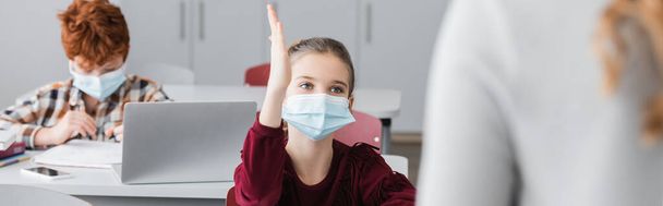 schoolmeisje in medisch masker rozijn hand tijdens les in de buurt van leraar op wazig voorgrond, banner - Foto, afbeelding