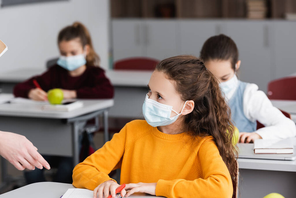 учительница указывает рукой на школьницу в медицинской маске в классе - Фото, изображение