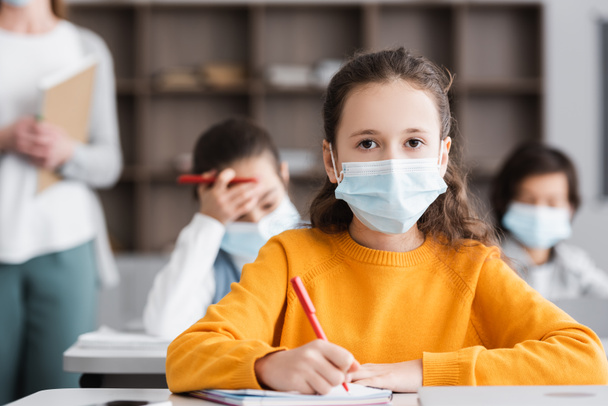 girl in medical mask looking at camera near teacher and classmates on blurred background - Photo, Image