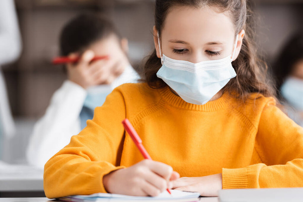 школьница в медицинской маске пишет в блокноте во время школьного урока - Фото, изображение